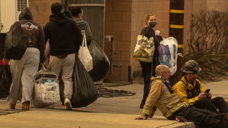 人々が所持品を手に、休息する消防士のそばを通り過ぎる＝アルタデナ/David McNew/Getty Images