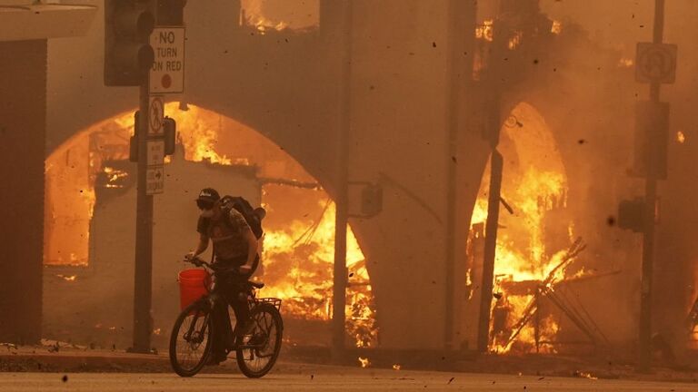 燃える建物を横目に自転車をこぐ人＝アルタデナ/Chris Pizzello/AP